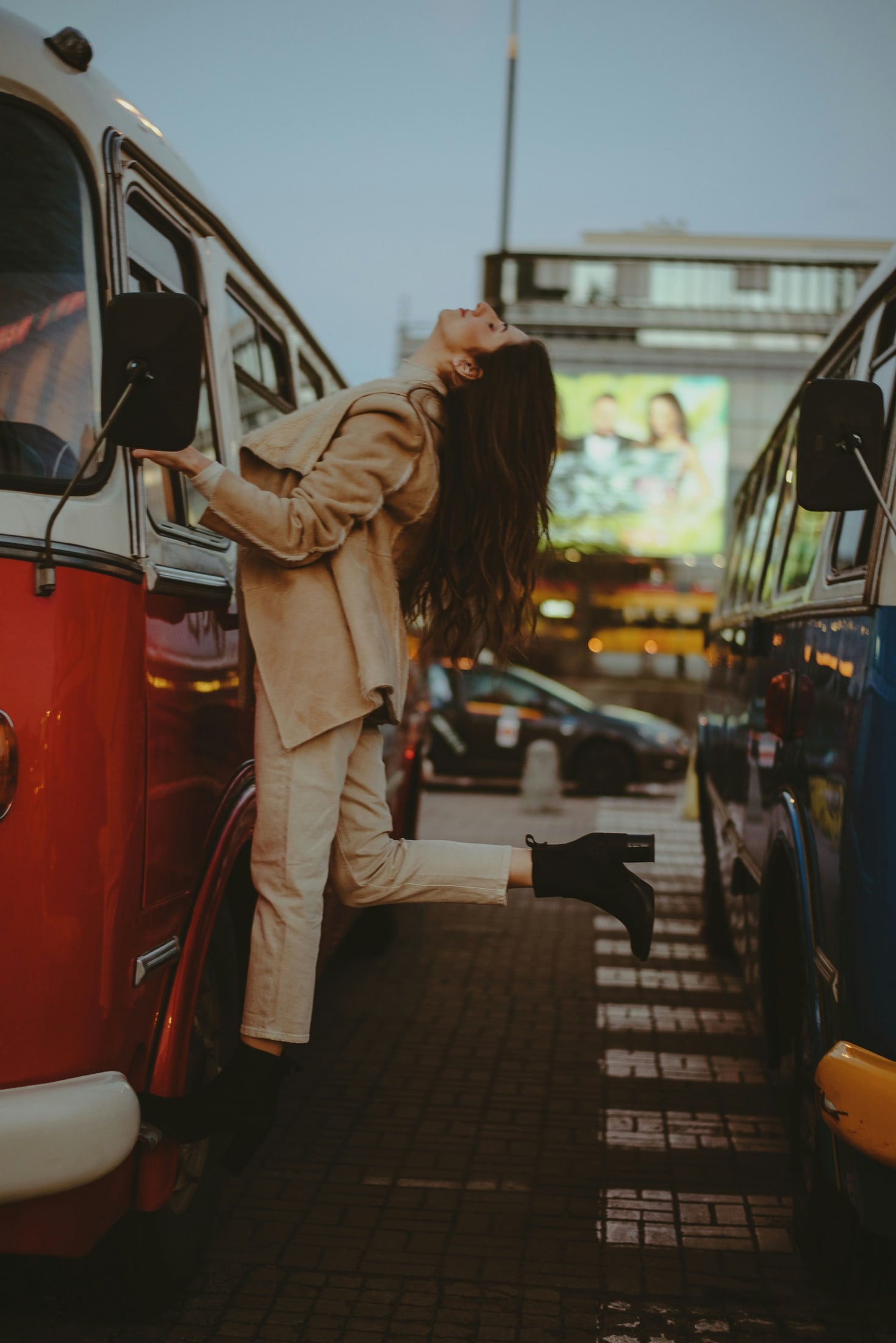 A woman leans back from the window of a red vintage bus at dusk, one foot kicked up.