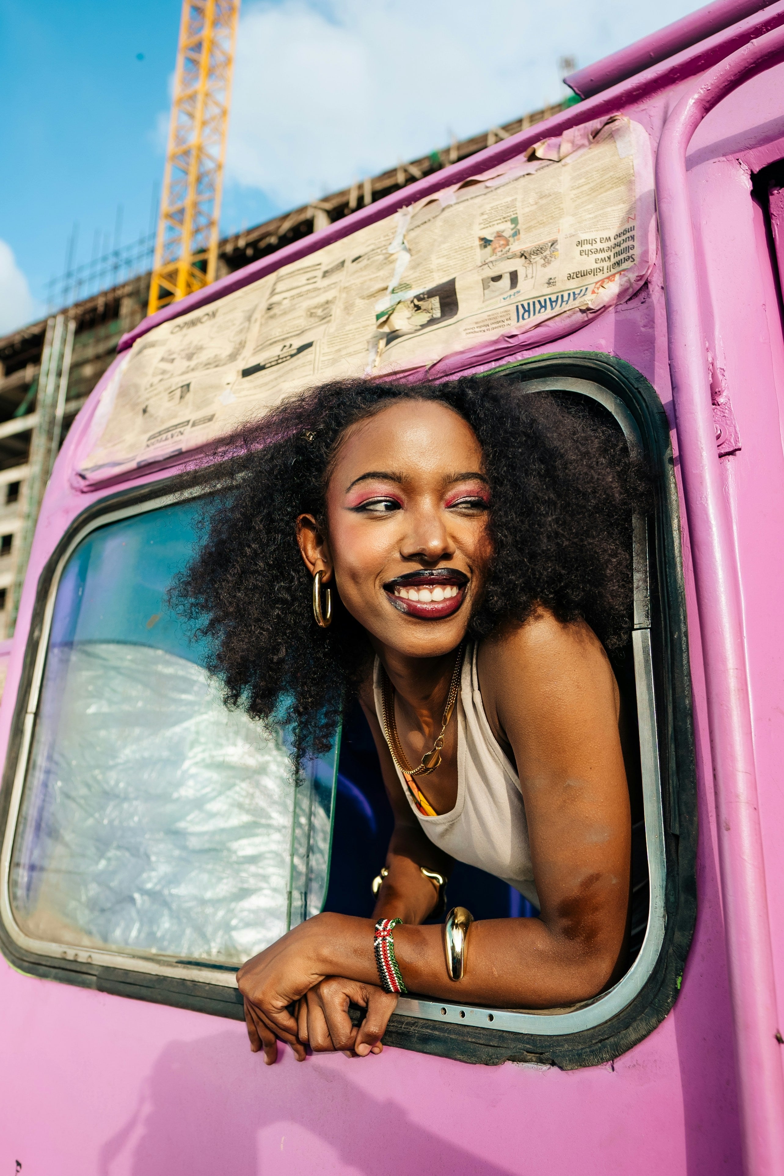 A woman leans out of the window of a pink minibus, smiling at something outside.