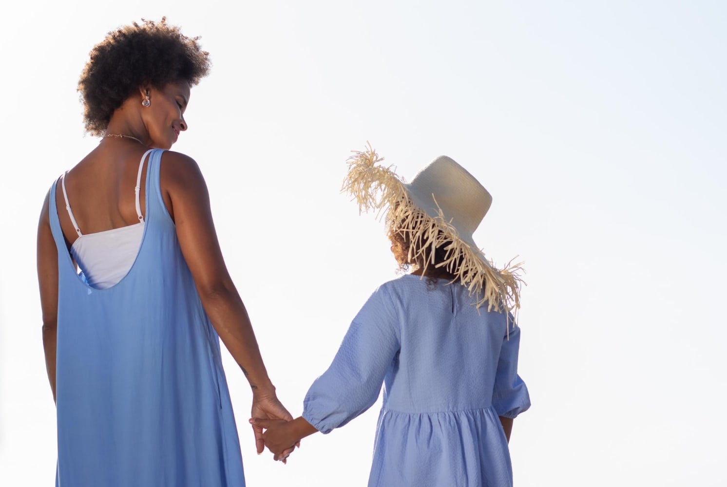 Seen from behind, a woman and a girl in blue dresses walk hand in hand under a pale sky.
